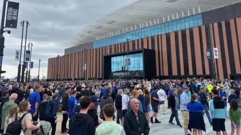 Everton fans stand on the plaza outside the Hill Dickinson stadium ahead of the match with Roma. There are thousands of fans, many wearing Everton shirts or other blue tops, in front of the bronze and black stripes that ring the stadium.