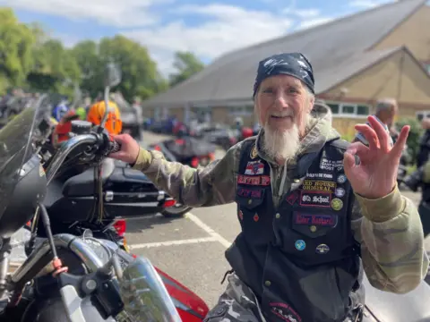 Jenny Kirk/BBC An older man with a long beard on a motorbike. He is wearing a bandana and a denim gilet with badges stitched on. He is smiling.