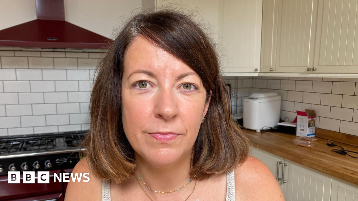 A woman with a brown bob. She is looking at the camera and is sitting in a kitchen, with a red stove in the background