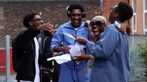 EPA A group of four friends open their exam results and another classmate looks on in the background. They are smiling and and have their arms around one another. The student on the far left wears thick rimmed glasses and a black hoodie over a white t-shirt, and is facing the student to his right, who is wearing headphones and a blue tracksuit. Second from the right, another student with short blonde hair and sunglasses smiles towards the camera. Furthest on the right, a student in a blue shirt wearing glasses looks away from the camera towards the rest of the group.