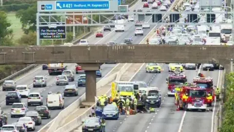 National Highways The M1 motorway, showing eight lanes of traffic, with vehicles on the road, and cars stopped. There are fire engines, ambulances and police vehicles on several lanes. 