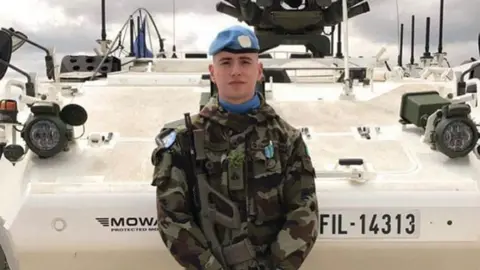 Irish Defence Forces Irish soldier Seán Rooney, wearing camouflage clothes and a blue beret. He is holding a gun and standing in front of an armoured vehicle.