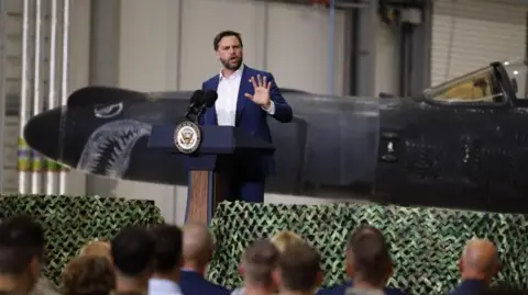 EPA A man with dark hair and a beard makes a speech standing in front of a military aircraft with a face painted on the nose. 