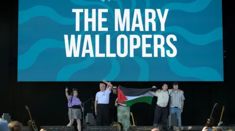 Getty Images Multiple members of The Mary Wallopers stood on the Victorious Festival stage holding a Palestinian flag and with mouths open and arms raised appearing to chant. Behind them is a large screen with their band name on.