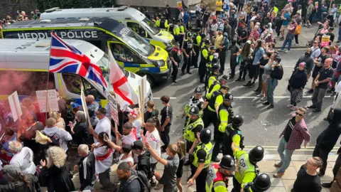Looking down on the two sets of protestors, a line of police in high-viz vests standing between them, large police vans are also present. They are blocking a road, and protestors on the left hold flags.