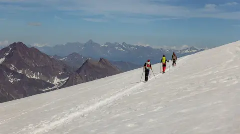 TOBY RONEY Three hikers with backpacks and poles trekking along a snow-covered mountain trail, with rugged peaks and a partly cloudy sky in the background.