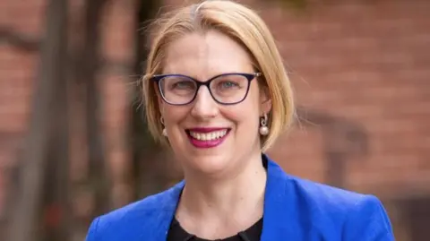 Supplied A blonde woman wearing a black shirt, blue blazer and eye glasses looks at the camera. She is wearing pearl earrings and lipstick
