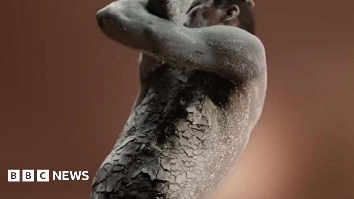 A topless man with black skin visible from his torso up covered in a grey clay-like material that is cracked. He is holding his arms across his face as though he is in pain.