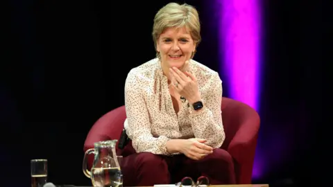 PA Media Nicola Sturgeon smiling straight at the camera. She is wearing a white shirt with a pattern on it. She is wearing maroon trousers. She is sitting on a dark red chair. Her right hand is raised under her chin while her left arm is across her lap. A jug of water and a glas off water and a pair of glasses are on a table in front of her. The background is dark with a beam of purple light coming from behind the left side of the chair.