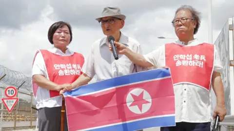 Jungmin Choi/BBC Ahn Hak-sop, wearing glasses, a hat, and a white shirt, holds a North Korean flag while two volunteers in red vests stand beside him - one holding a microphone for him, the other holding his right hand.