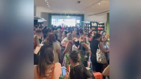 Kellie Greenhalgh A picture inside a large, busy bookshop, with groups of people stretching back towards the far, windowed wall.
