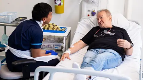 PA Media A hospital room with a male patient lying on a bed and a healthcare worker in scrubs attending to the patient's arm. The patient is dressed in blue jeans and a black t-shirt, and appears to be receiving medical attention. Medical equipment, including a trolley with medication and supplies, are visible beside the bed.