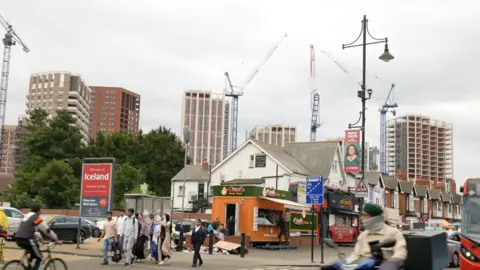 A street corner with a group of people crossing the road and people riding bikes and mopeds. Behind the junction is a row of houses with a car park for an Iceland shop and then cranes and several newly-built tower blocks can be seen in the background