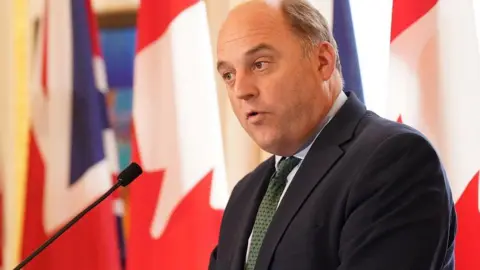 PA Media Sir Ben Wallace in a dark suit and tie gives a speech against the backdrop of union flags
