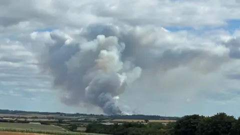 White plume of smoke swirling into the sky, fields a seen in the foreground.