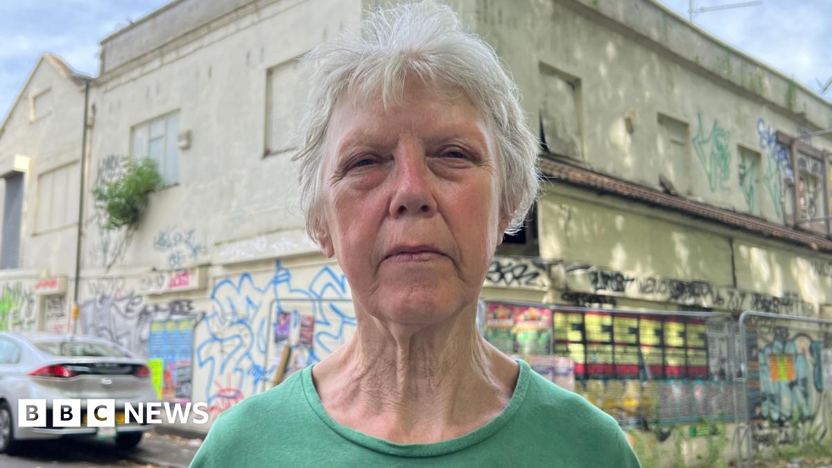 Ann Devereaux stands beside the derelict property next door - a large building now overtaken by decay. Graffiti covers the walls, weeds and shrubs grow unchecked from cracks in the brickwork, and gaping holes expose the interior to the elements.