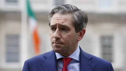 PA Media Tánaiste Simon Harris, pictured from the chest up, wearing a navy suit, light blue shirt and red tie. He has greying short hair. There is a blurred grey building and Irish flag in the background. 