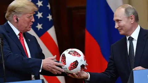 Getty Images Vladimir Putin smiles as he hands a white and red football to Donald Trump during a meeting in Helsinki, Finland, in July 2018. Behind the pair are a US and a Russian flag. 