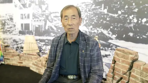 BBC/Hyojung Kim An old man in a chequered blazer sits in front of a black-and-white photograph showing destroyed streets and debris 