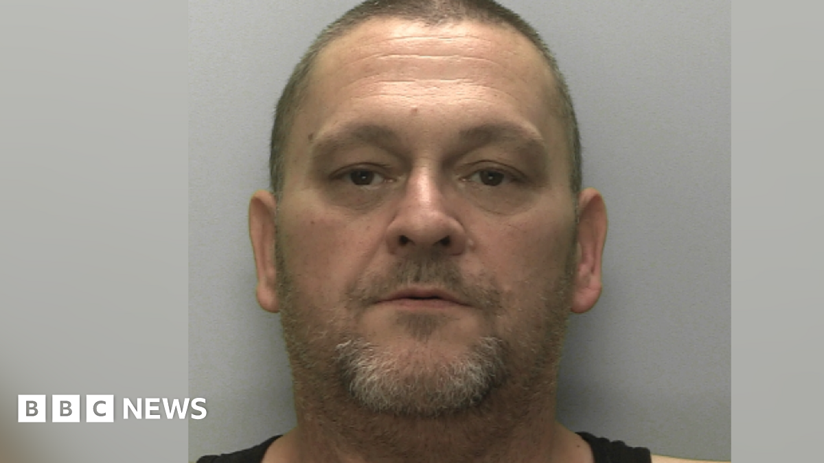 A man, with a small beard and brown eyes, looking directly at the camera. There is a grey wall behind him.