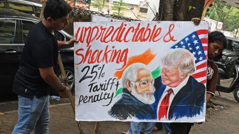 Ashish Vaishnav/SOPA Images/LightRocket via Getty Images Two young people carry a handpainted poster of Prime Minister of India Narendra Modi and U.S. President Donald Trump with the writing "Unpredictable & shocking 25% tariff + penalty"