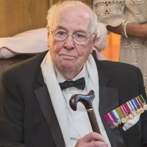 Berry Vissers/Squadron Prints An older John Cruickshank in recent years. He is looking slightly off camera. The top of his head is bald with white hair round the side. He is wearing light-framed glasses and a black suit jacket over a white shirt with a black bow tie. He has a collection of colourful medals on his upper left chest and is holding a walking stick in his right hand.
