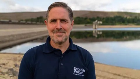 Brian McCarthy standing in front of a resrvoir. he has short brown hair and a beard and is wearing a dark blue polo shirt with a Scottish water logo