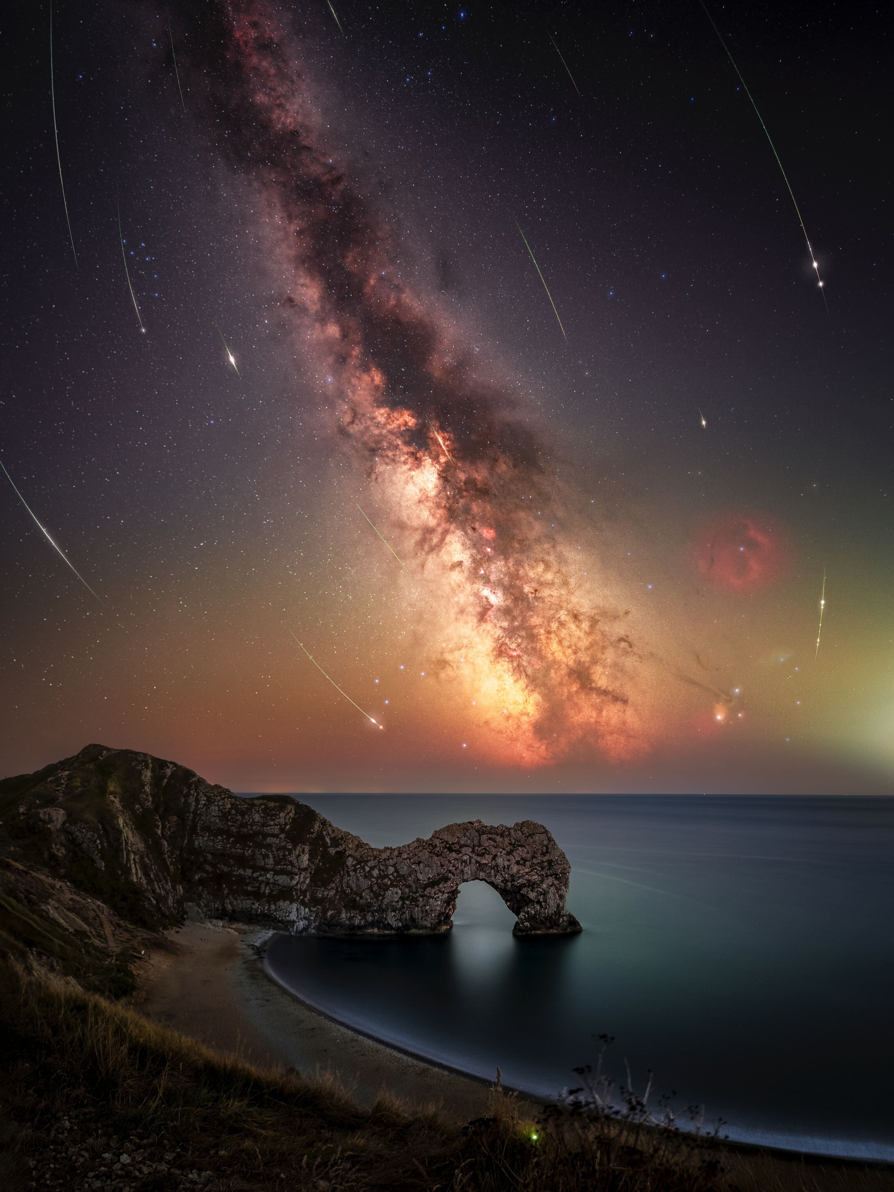 Perseid meteors are pictured steaking through the sky next to the glowing band of the Milky Way, arcing downwards towards a coastal scene featuring an arch-like rock formation.
