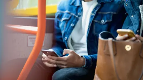 SolStock/Getty Images A man in a white t-shirt and blue denim jacket sits on a bus next to the window and uses his phone. His face is out of the camera shot. Another passenger sat next to him also uses their phone.