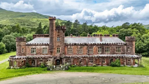 Nest Marketing An exterior view of the red sandstone lodge with its ornate turrets and battlement-like rooftops. The lodge is set in an grounds with large areas of grass and trees.
