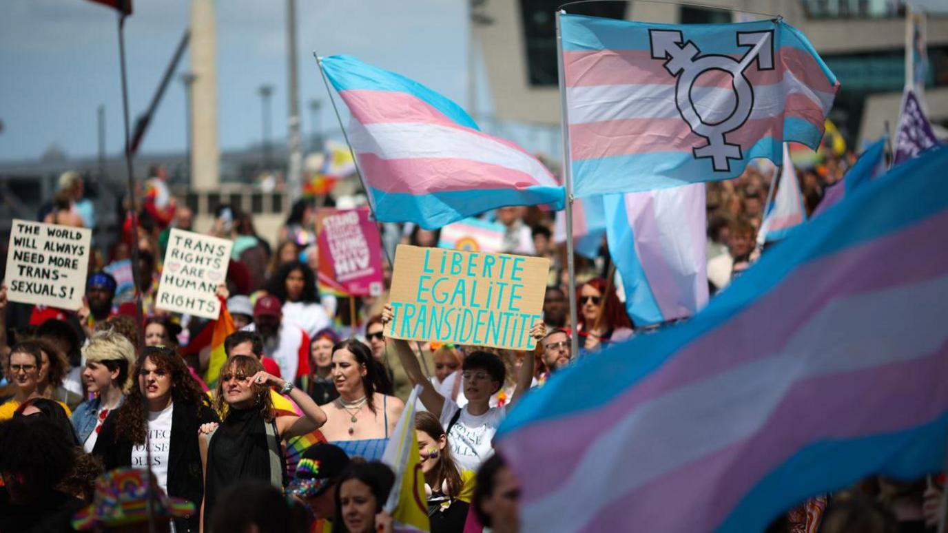 Marchers taking part in Liverpool Pride. Many in the crowd are holding placards supporting trans-rights while others are waving the transgender pride flag which consists of five stripes of blue, pink and white.
