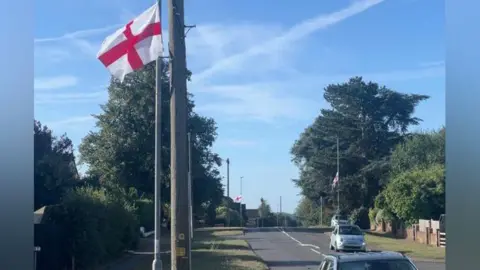 BBC A red and white St George's Flag is tied to a lamppost beside a residential road, and there are two more in the background. There are a couple cars in the frame. There is a blue sky.