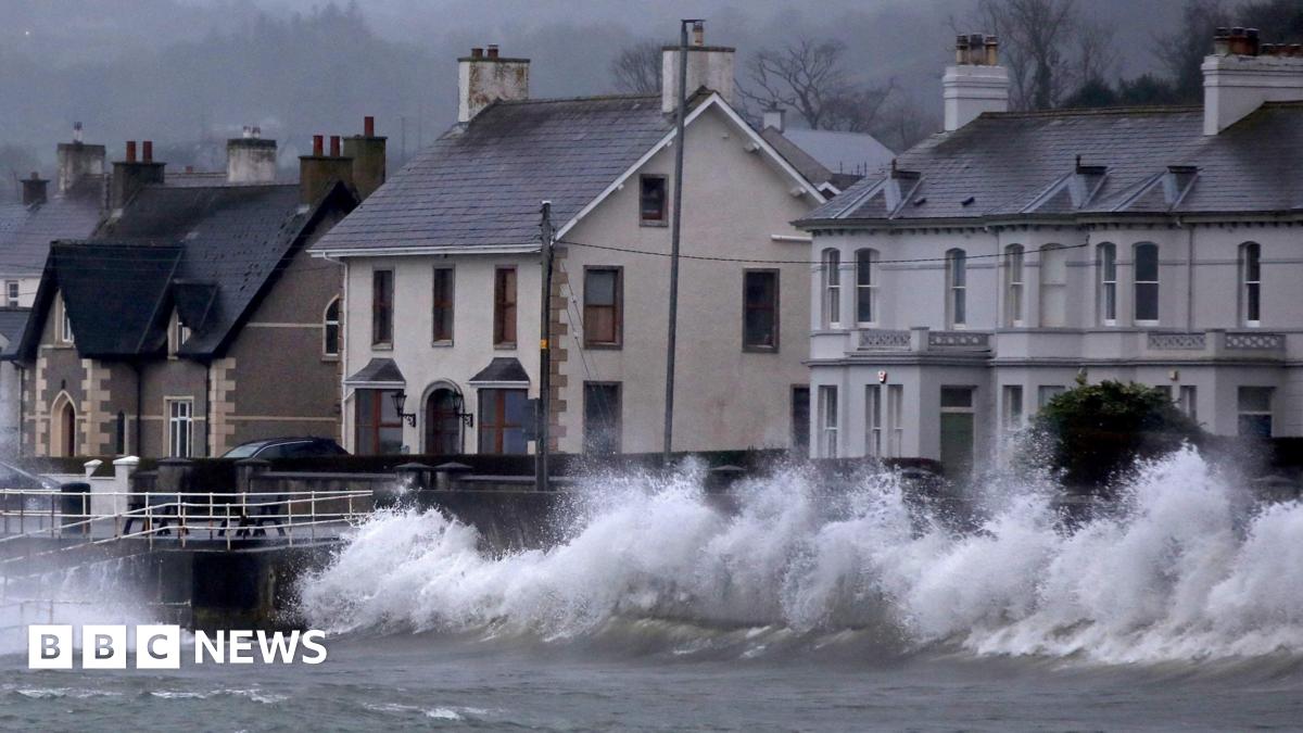 A row of houses by the sea. There are large waves hitting the wall between the sea and the houses.