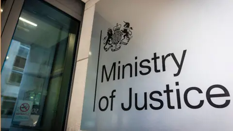 Getty Images A close-up view of the entrance to a Ministry of Justice building. The image shows a glass door on the left with a 'No Smoking' sign and a large frosted glass panel on the right with the text 'Ministry of Justice' and an official crest above it.