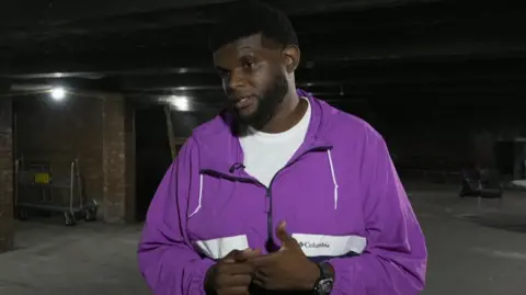 A man in a purple tracksuit with black hair and dark eyes stands in a storage space. He is being interviewed for TV and is wearing a radio mic.