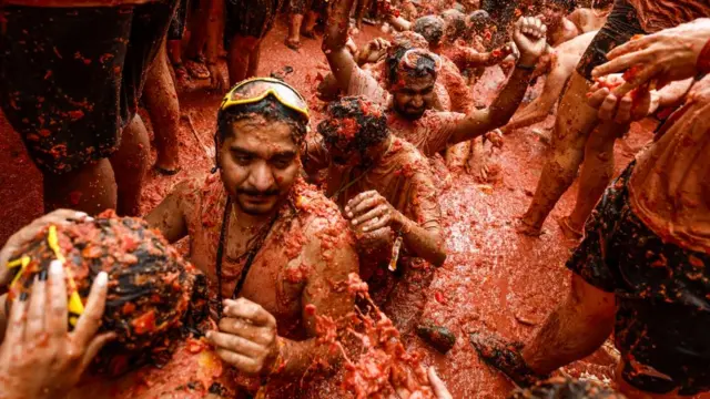 Flanked by crowds, a line of people sit in ankle-deep tomato pulp that covers the crowd, some wearing goggles