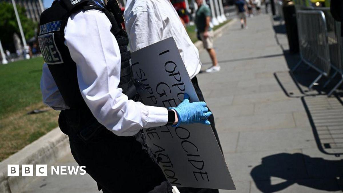 A close up shot of a female police officer's torso as she holds a sign that she has confiscated that reads: I oppose genocide, I support Palestine Action. She is wearing police uniform and blue gloves