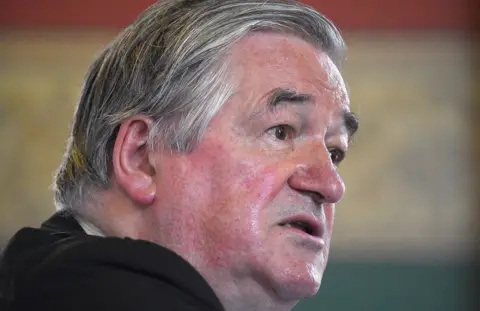 PA Media A close-up image of the side profile of a person with gray hair. The background is out of focus and appears to be indoors with a mix of red, green, and beige colors