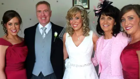 Rodgers family Tony Rodgers with his wife and daughters on one of their wedding days. Tony has short grey hair and is wearing a formal wedding suit with a black suit jacket over a grey waistcoat, silver tie and white shirt. One of his daughters has blonde hair and is wearing a white wedding dress. Two of the others at each side of the photo have short brown hair and are wearing maroon bridesmaid dresses. His wife, second from right, has shoulder-length dark brown hair and is wearing a pink dress and a pink fascinator.