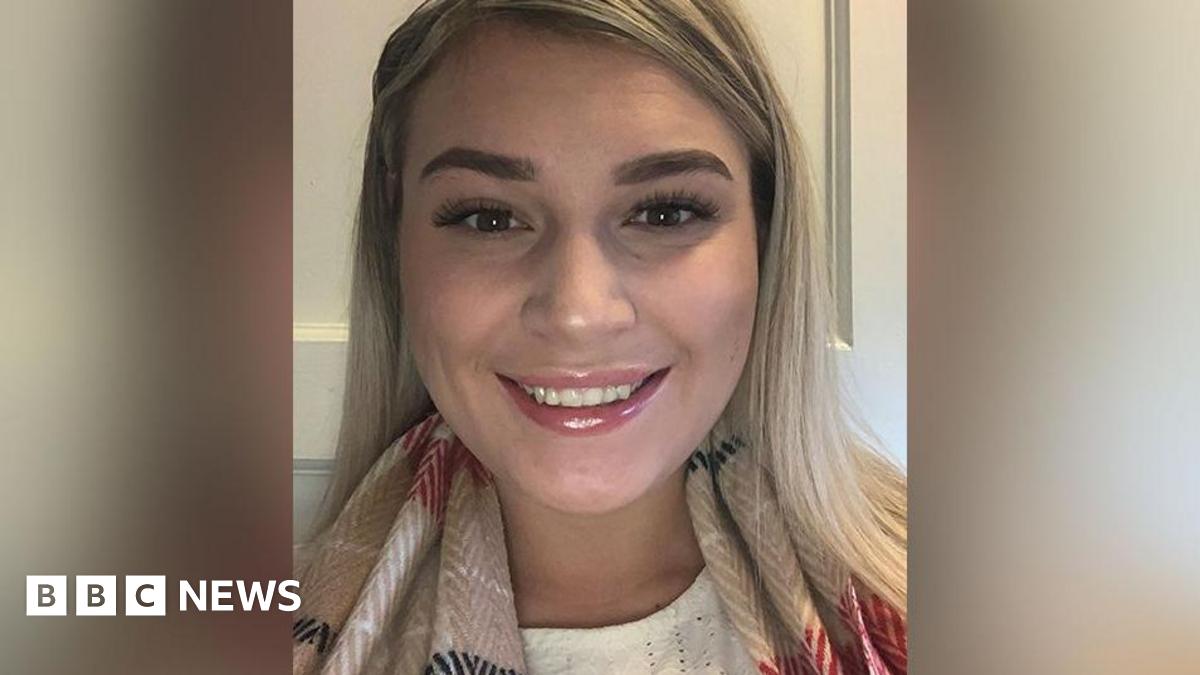 Close up of Leonie Wilkinson who has blond hair, brown eyes and brown eyebrows wearing a cream top and beige scarf with red, white and black patterns standing in front of a white cupboard. She is smiling.