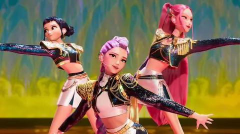 Netflix A scene from the animation Kpop Demon Hunters, where three young women - K-pop idols - are dressed in shiny costumes and performing on a stage. They are posing with their arms outstretched.
