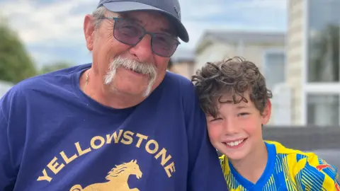 BBC Trevor Parry wearing a baseball cap and sunglasses smiles at the camera sat beside Elliott who has curly brown hair and is wearing a Stockport County football shirt.