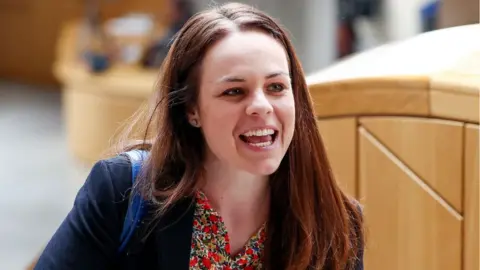 Getty Images Kate Forbes, who has brown hair, is smiling in the Scottish Parliament. She is wearing a dark blue jacket and red floral shirt. 