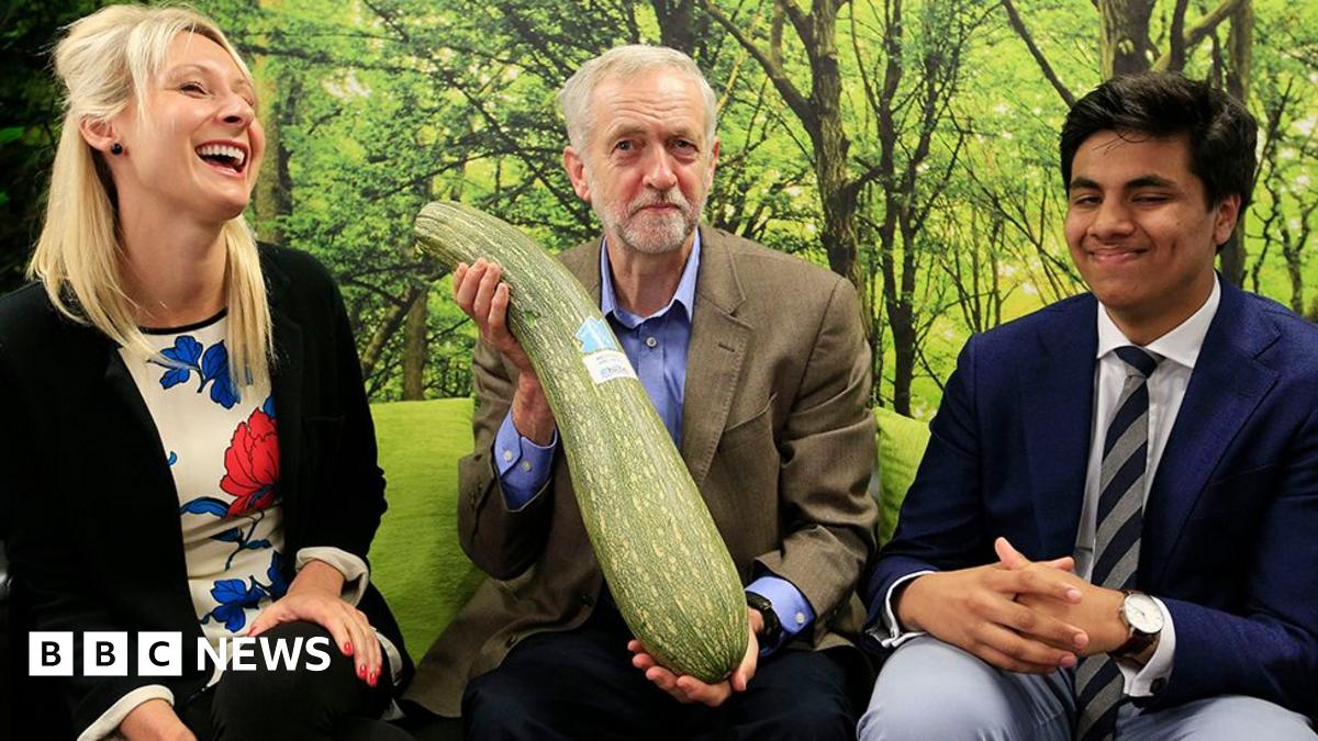 Jeremy Corbyn holds a large marrow with one eyebrow raised, flanked by a young woman who is laughing and a young man in suit and tie, who is smiling