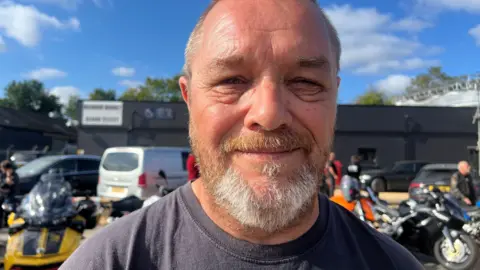Jenny Kirk/BBC A man with a beard looking at the camera and smiling. There are motorbikes parked behind him.