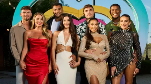ITV/Shutterstock The eight Love Island finalists stood smiling in front of a blue, pink and yellow heart. 