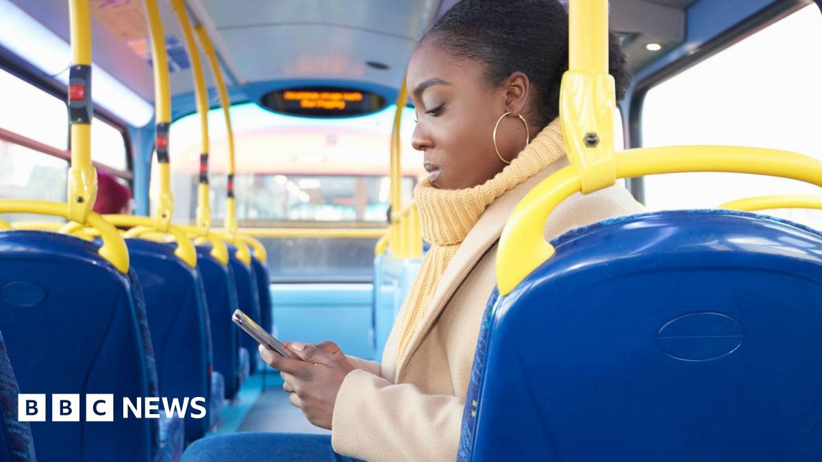 A young woman with her hair tied back sitting sideways on a bus looking down at her smartphone