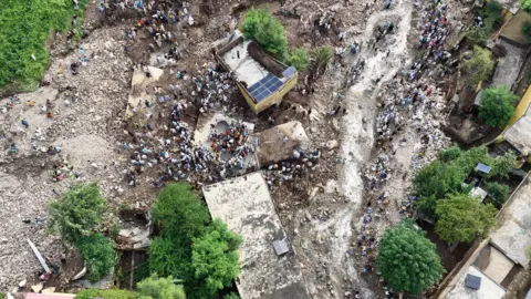 Overhead shot of flood damage with people watching. There are pockets of green which seem to be trees or banks, with mud running in between