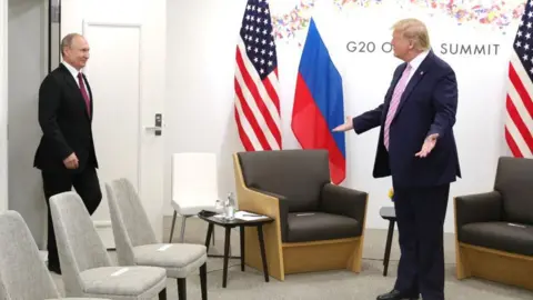 Getty Images Donald Trump, standing with his hands open at his sides, greets Vladimir Putin in a white room lined with US and Russian flags in a meeting at the G20 summit in Osaka, Japan, in June 2019