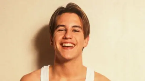 Getty Images Anthony Kavanagh as a young star grins for a photo shoot. He has light coloured hair and is wearing a white vest top.